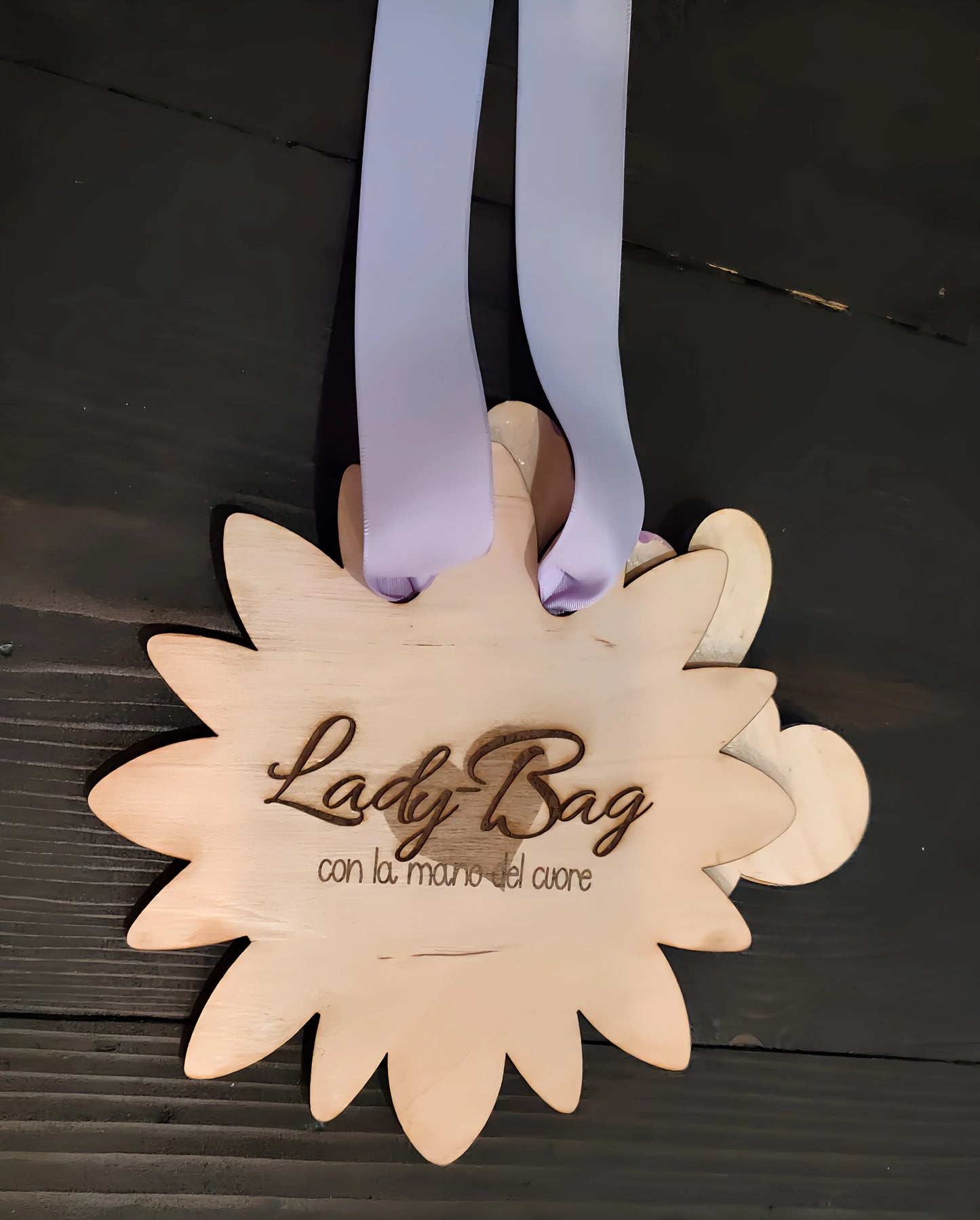 Wooden flower-shaped plaque with 'Lady Bag' engraved, lilac ribbon, on dark wood table