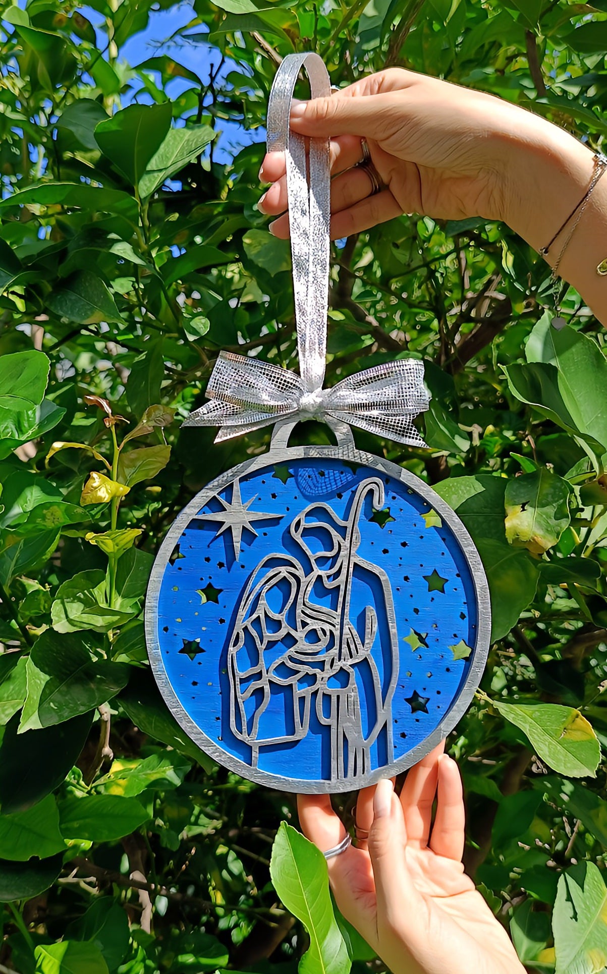 Hand holding Nativity ornament with blue background and silver bow in green garden