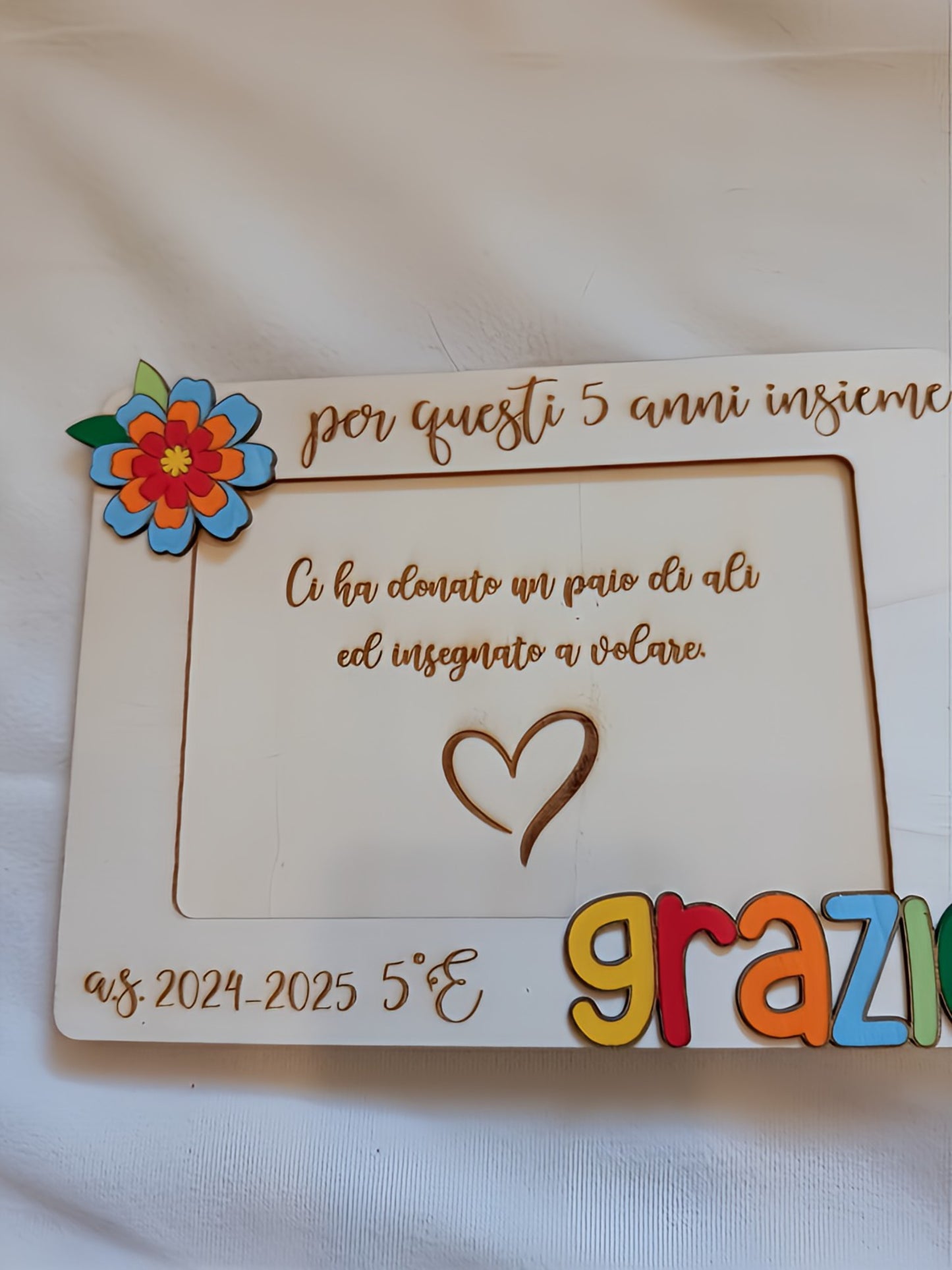 Vista frontale del portafoto in legno per maestra con cornice bianca, apertura centrale per foto 10x15, incisioni a pirografia con dedica personalizzata, scritta "grazie" in lettere tridimensionali multicolori e fiore decorativo in legno dipinto nell'angolo.
