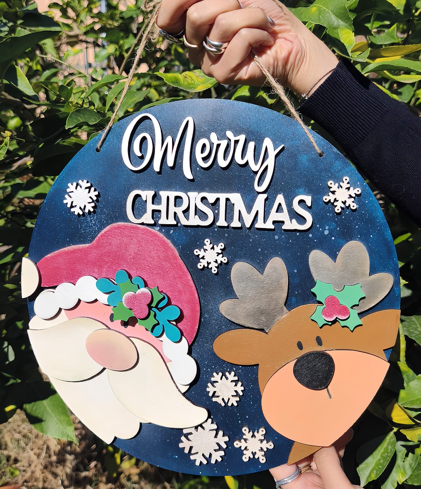 Hand holding round Merry Christmas sign with Santa, reindeer, snowflakes, greenery background