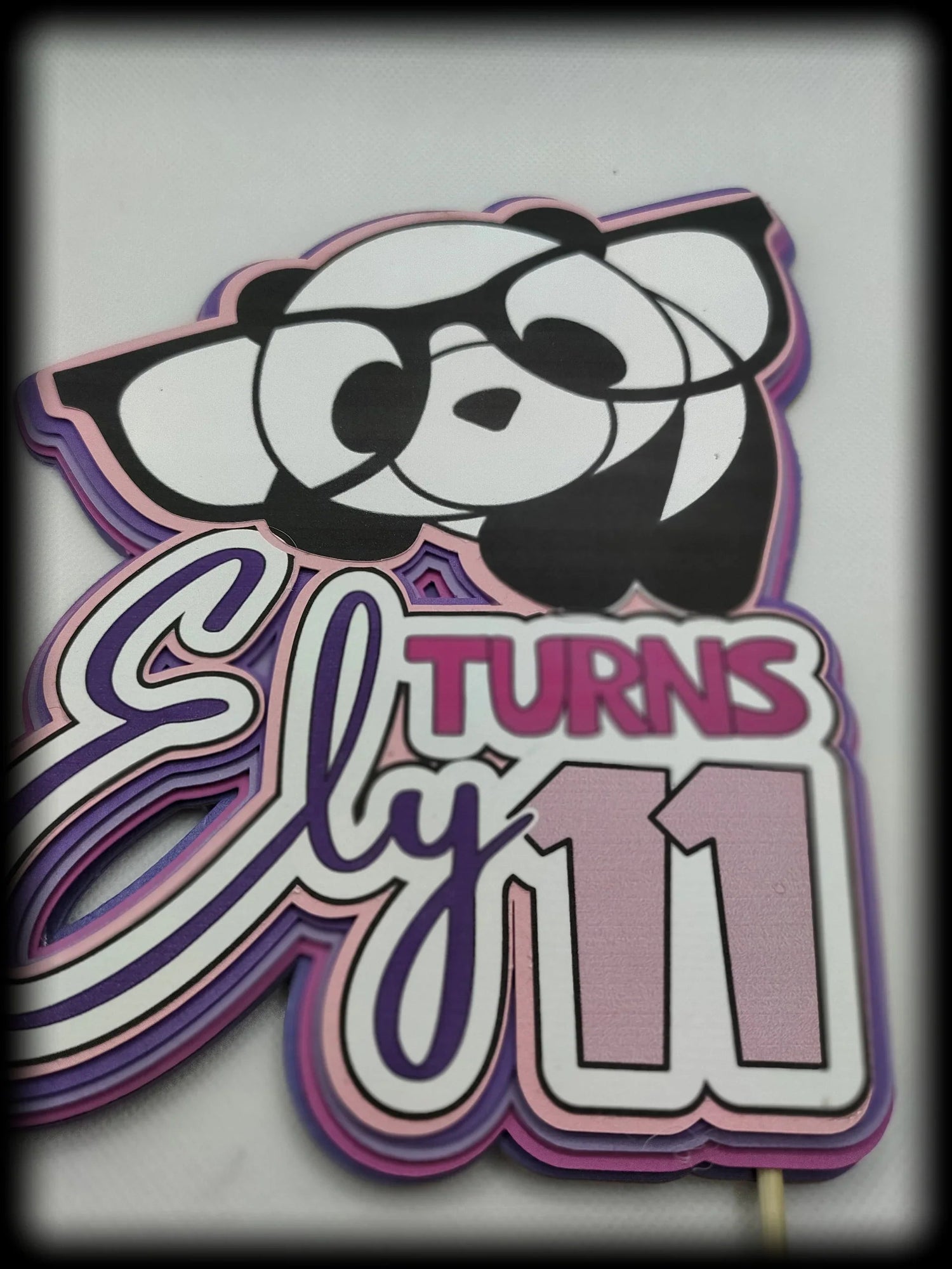 Birthday cake topper with cartoon panda and text 'Ely turns 11' in pink and purple.