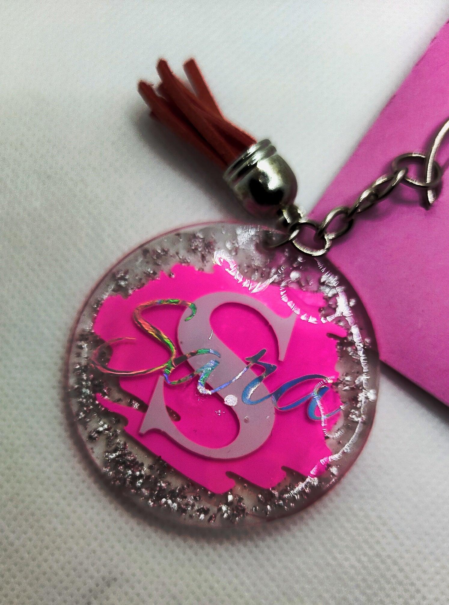 Personalized pink resin keychain with silver glitter, name Sara, and tassel on white fabric.