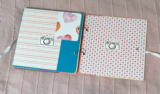 Cute scrapbook open to two pages with heart patterns and camera illustrations on a pink surface