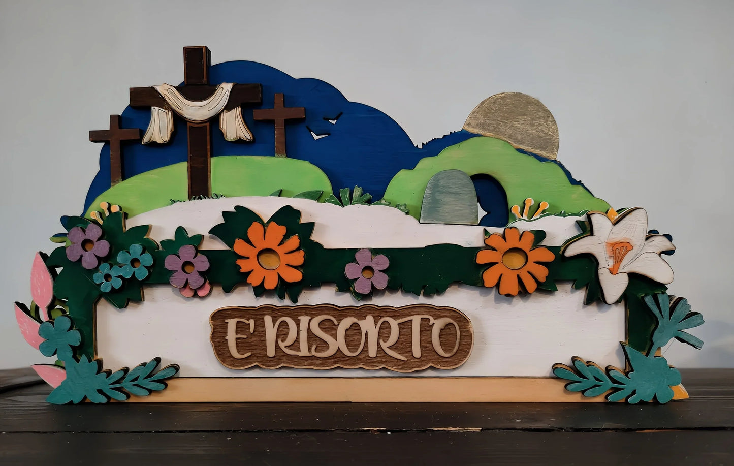 Wooden Easter resurrection scene with three crosses, flowers, and sign reading 'È risorto'.