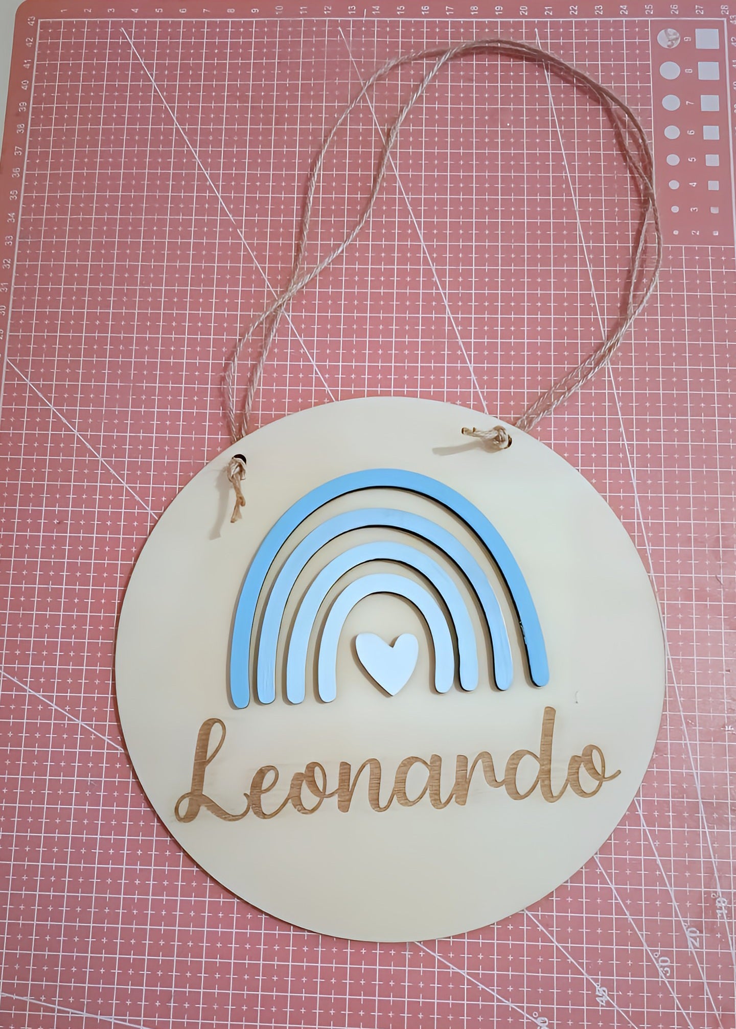 Round wooden name sign with blue rainbow, heart, and 'Leonardo' on a pink cutting mat