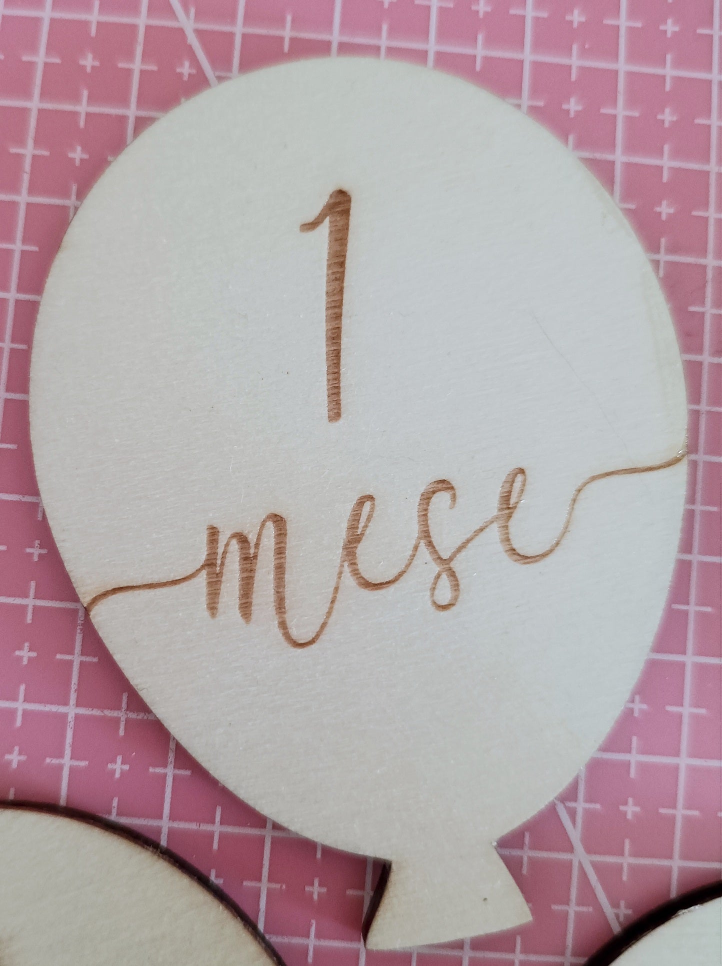 Wooden balloon-shaped plaque engraved with '1 mese' on pink grid background