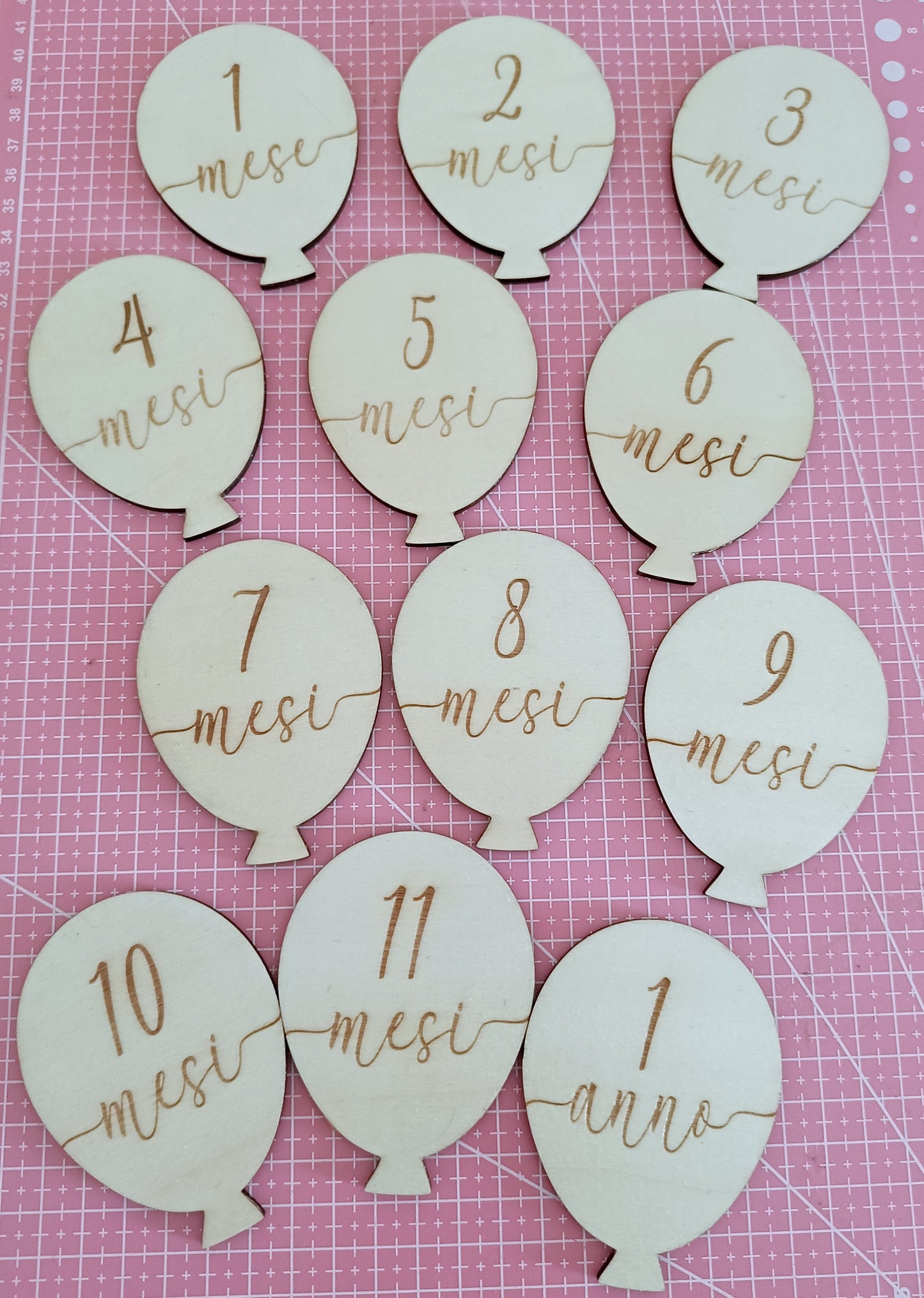 Wooden balloon-shaped milestone markers with Italian months and year on pink grid background