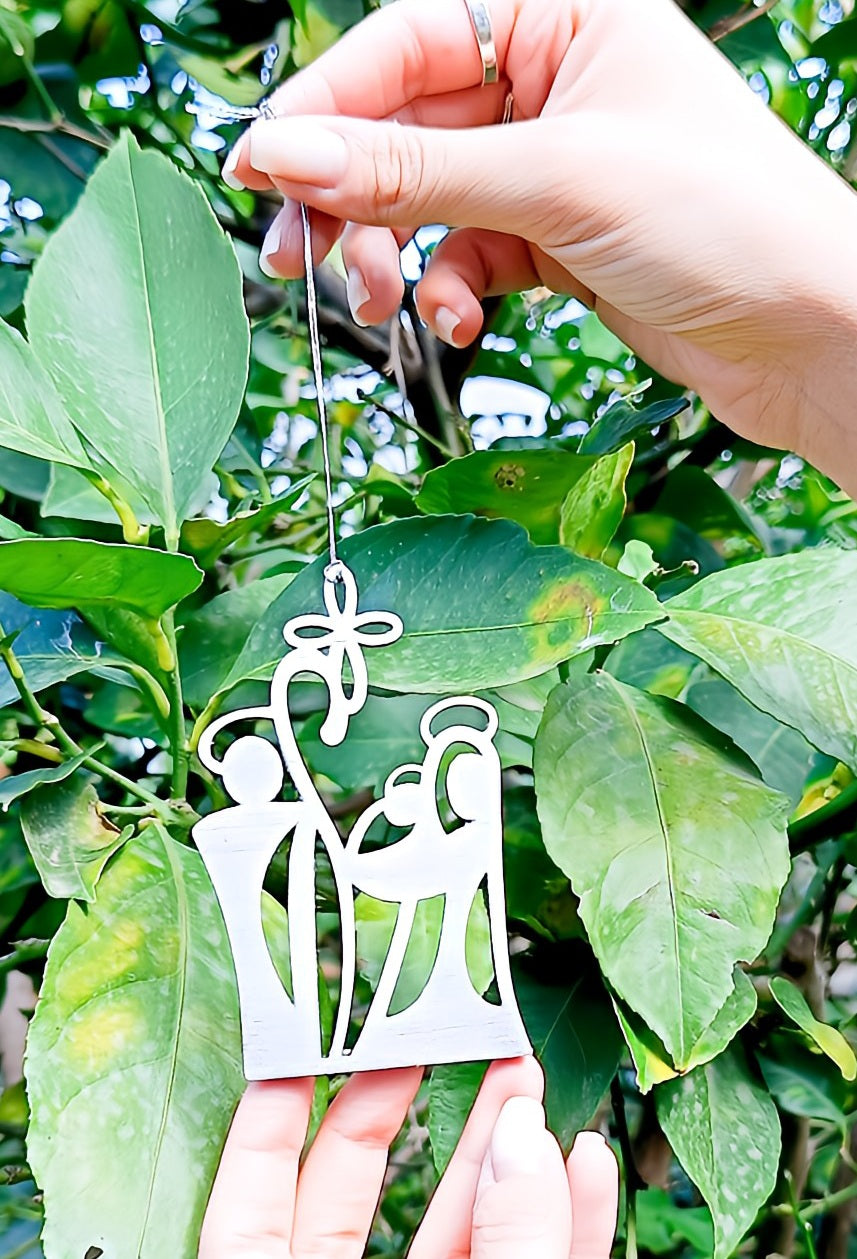 Hand holding metal nativity ornament near green leaves outdoors