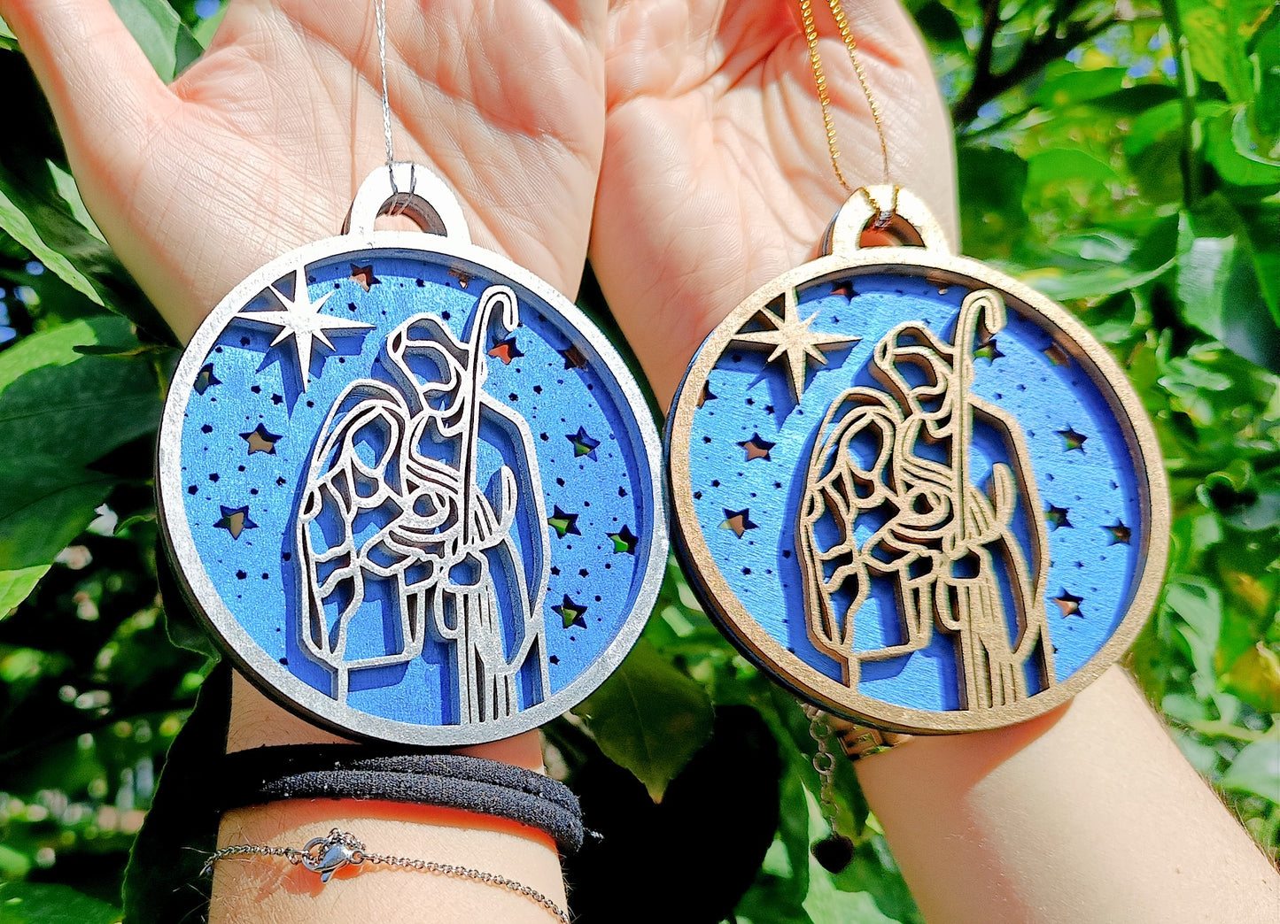 Hands holding two wooden Nativity Christmas ornaments with blue backgrounds outdoors