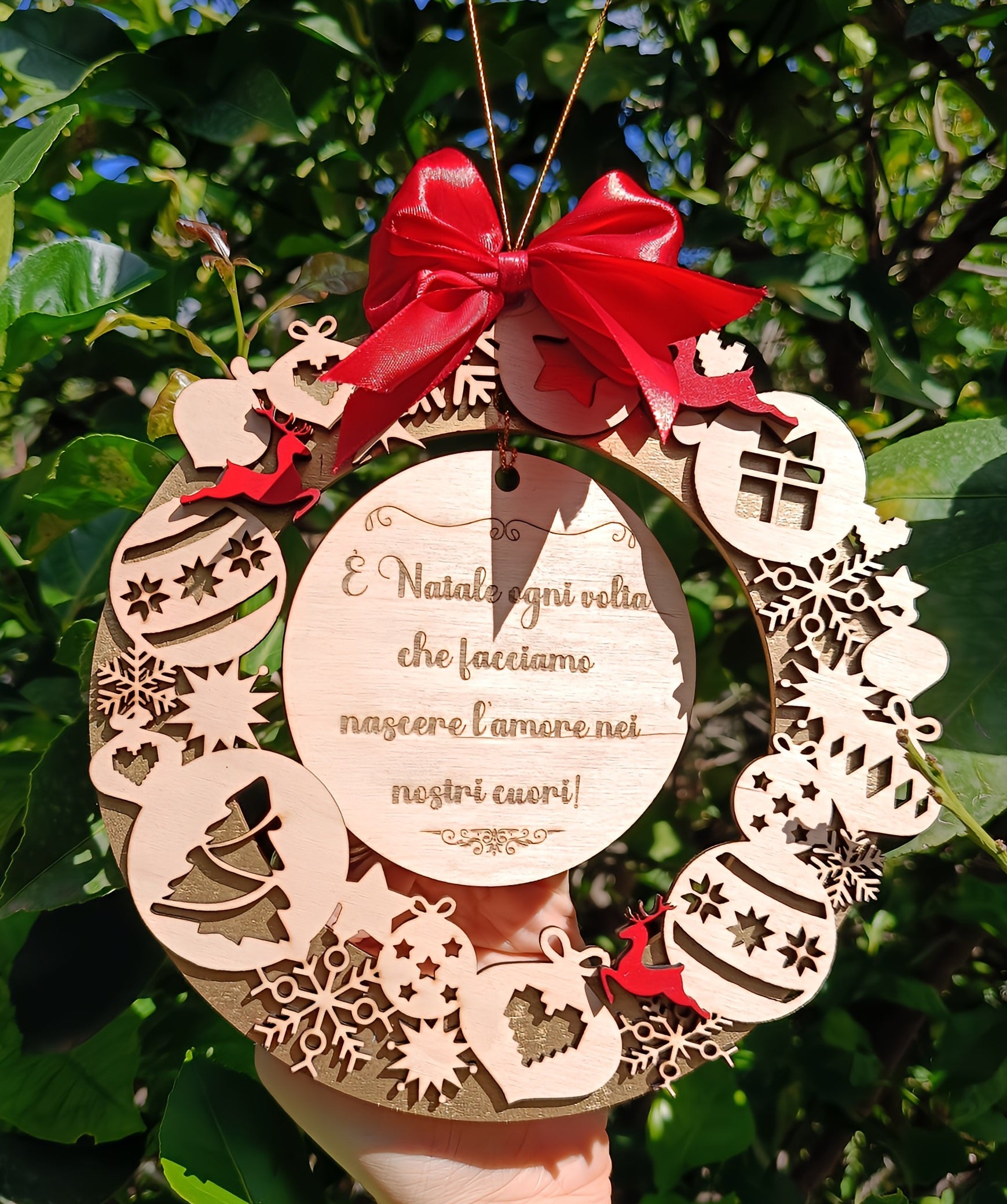 Hand holding wooden Christmas wreath ornament with red bow and Italian holiday message, outdoors