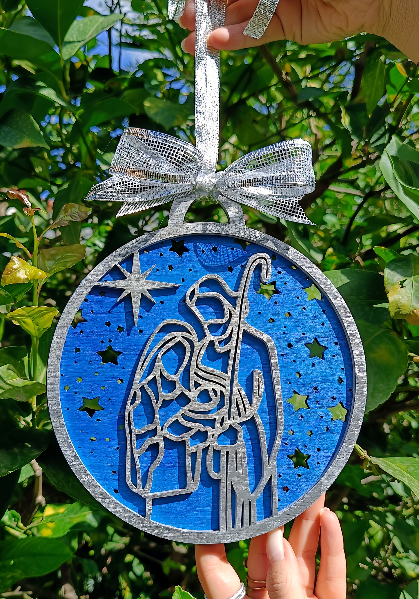 Hand holding blue nativity Christmas ornament with silver bow in garden setting