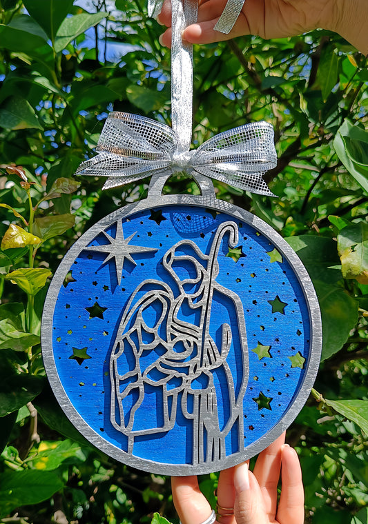 Hand holding blue nativity Christmas ornament with silver bow in garden setting