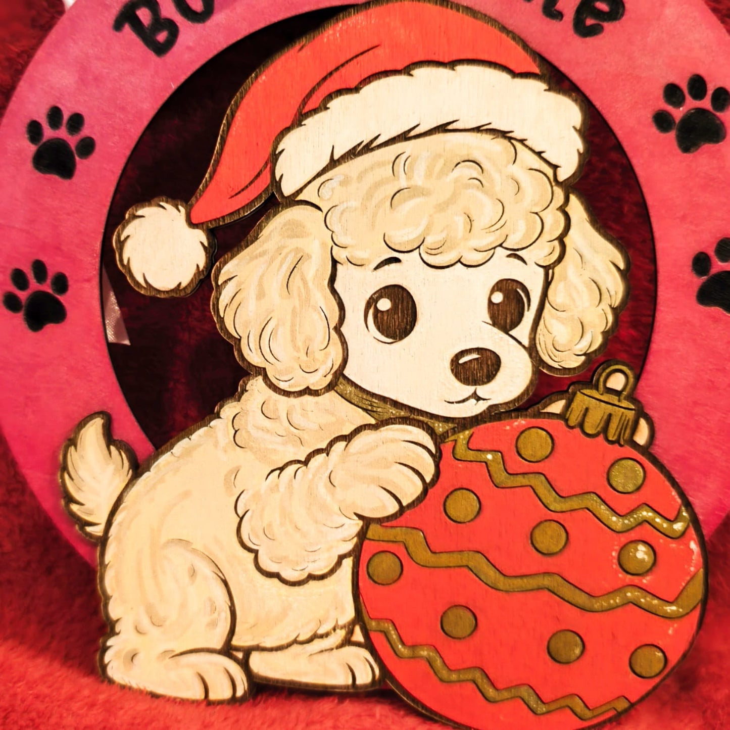 
Decorazione natalizia in legno con barboncino bianco che indossa cappello di Babbo Natale e tiene un ornamento rosso decorato. Scritta Buon Natale con impronte di zampe su sfondo rosa. Addobbo artigianale fatto a mano.