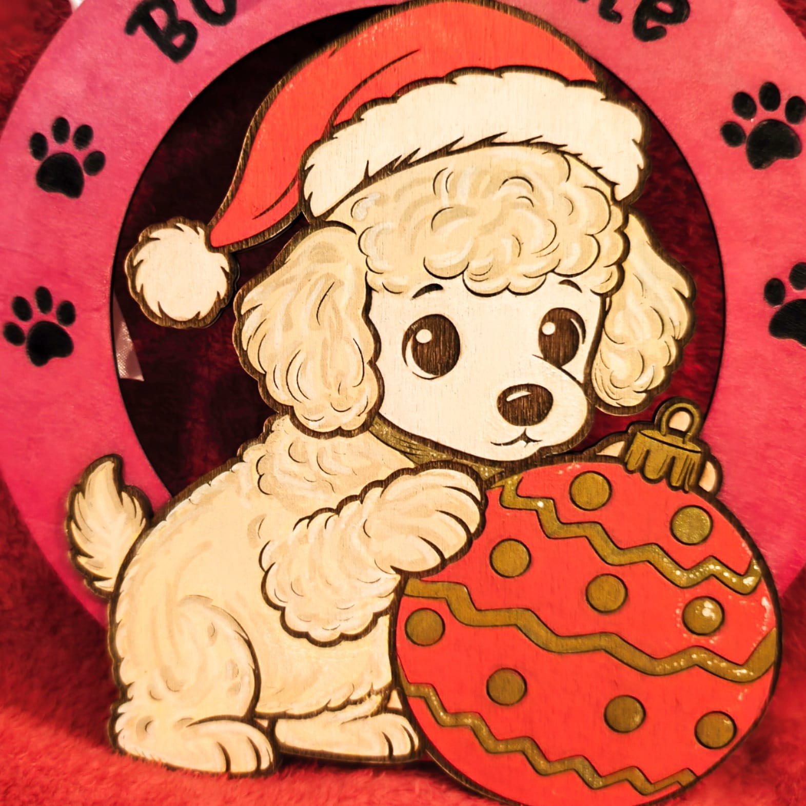 
Decorazione natalizia in legno con barboncino bianco che indossa cappello di Babbo Natale e tiene un ornamento rosso decorato. Scritta Buon Natale con impronte di zampe su sfondo rosa. Addobbo artigianale fatto a mano.