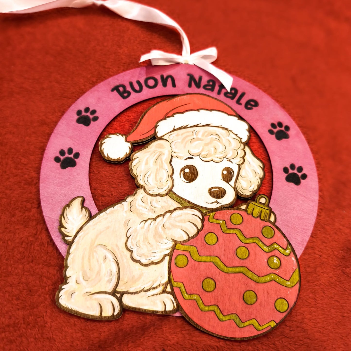 Addobbo natalizio artigianale in legno con barboncino bianco, cappello rosso di Babbo Natale e palla di Natale decorata. Scritta Buon Natale su cornice rosa con impronte di zampe. Nastro bianco per appendere. Decorazione handmade italiana.
