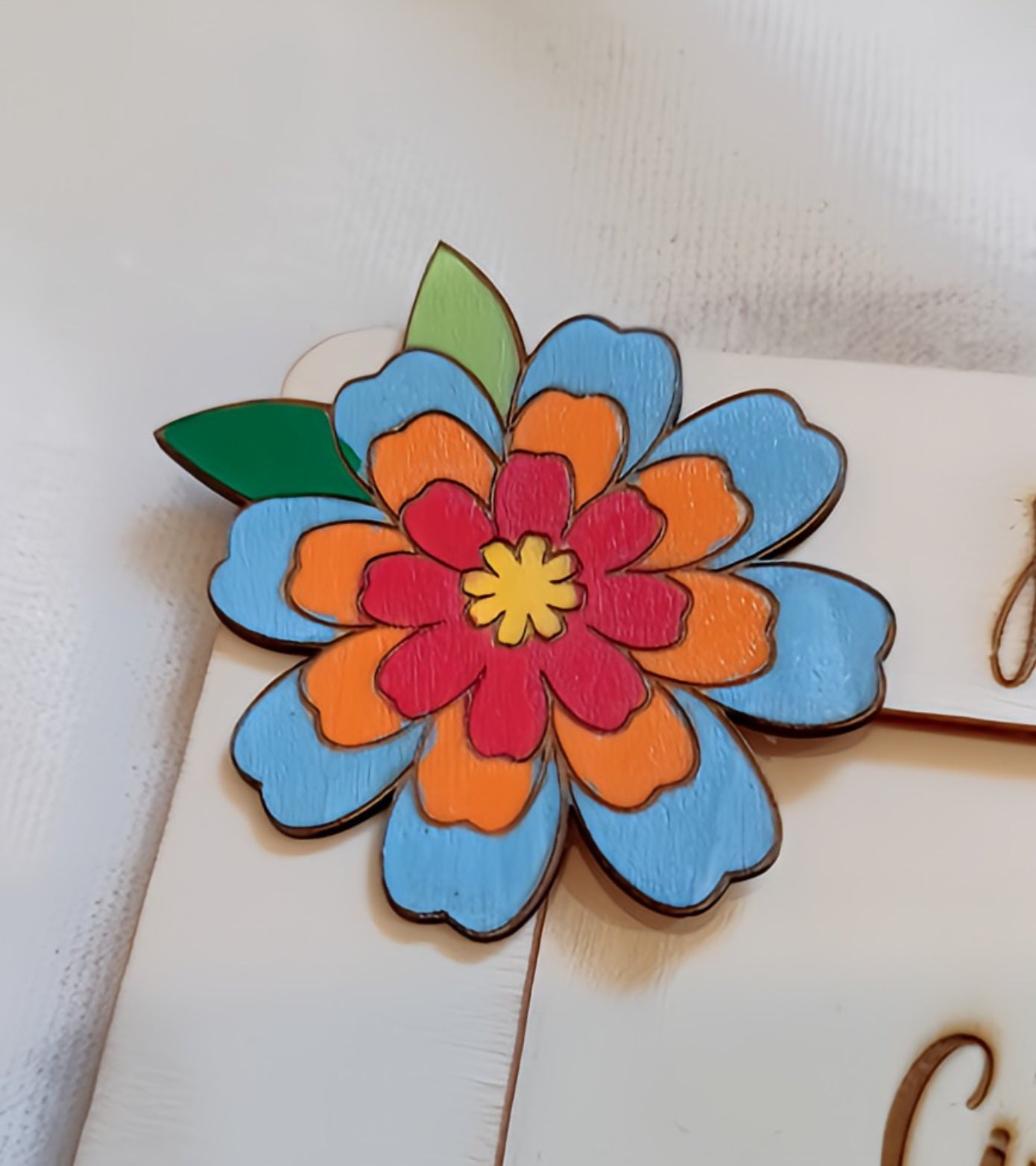 Dettaglio ravvicinato del fiore decorativo multistrato in legno dipinto a mano, con petali azzurri sul livello esterno, arancio sul livello intermedio, rosso-fucsia sul livello interno, centro giallo e foglie verdi. Elemento decorativo fatto a mano per portafoto regalo maestra.