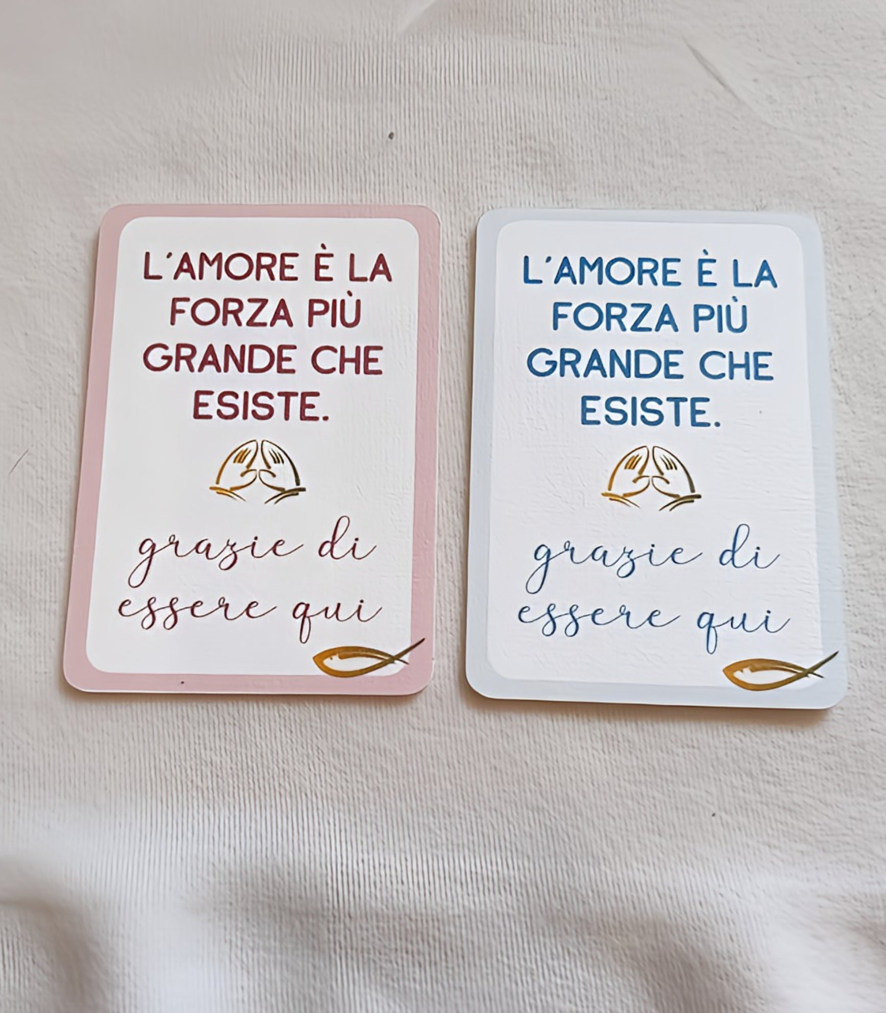 Two Italian gratitude cards with prayer hands, one pink and one blue, on a light fabric surface.