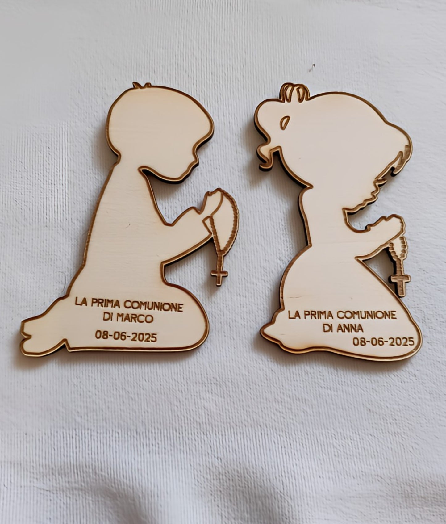 Wooden First Communion keepsakes shaped as praying boy and girl with rosaries on white fabric.
