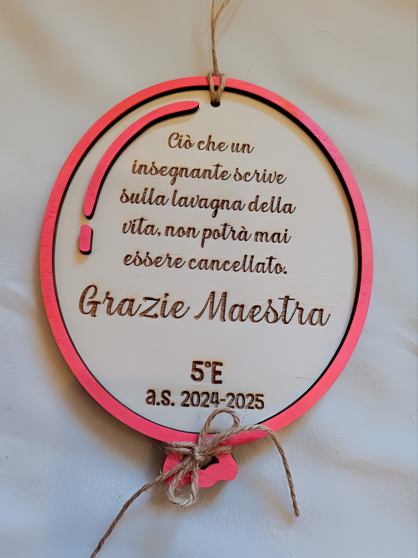 Targa rotonda in legno bianco con cornice multistrato fucsia-corallo, matita decorativa in legno rosa sul lato sinistro, incisione a pirografia della frase "Ciò che un insegnante scrive sulla lavagna della vita, non potrà mai essere cancellato. Grazie Maestra", personalizzazione 5°E a.s. 2024-2025, fiocco in cordino di iuta e cordino per appendere. Regalo di fine anno fatto a mano per insegnante.