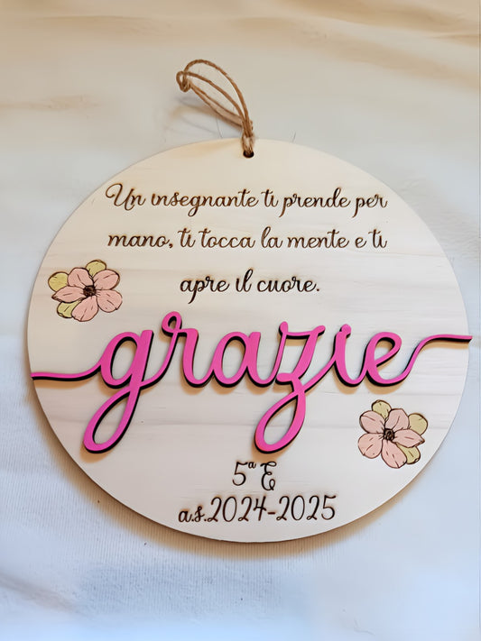 Targa rotonda in legno naturale con incisione a fuoco della frase "Un insegnante ti prende per mano, ti tocca la mente e ti apre il cuore", scritta tridimensionale "grazie" in legno dipinto fucsia, fiori decorativi dipinti a mano in rosa e verde, personalizzazione classe 5ª E anno scolastico 2024-2025 e cordino in iuta.