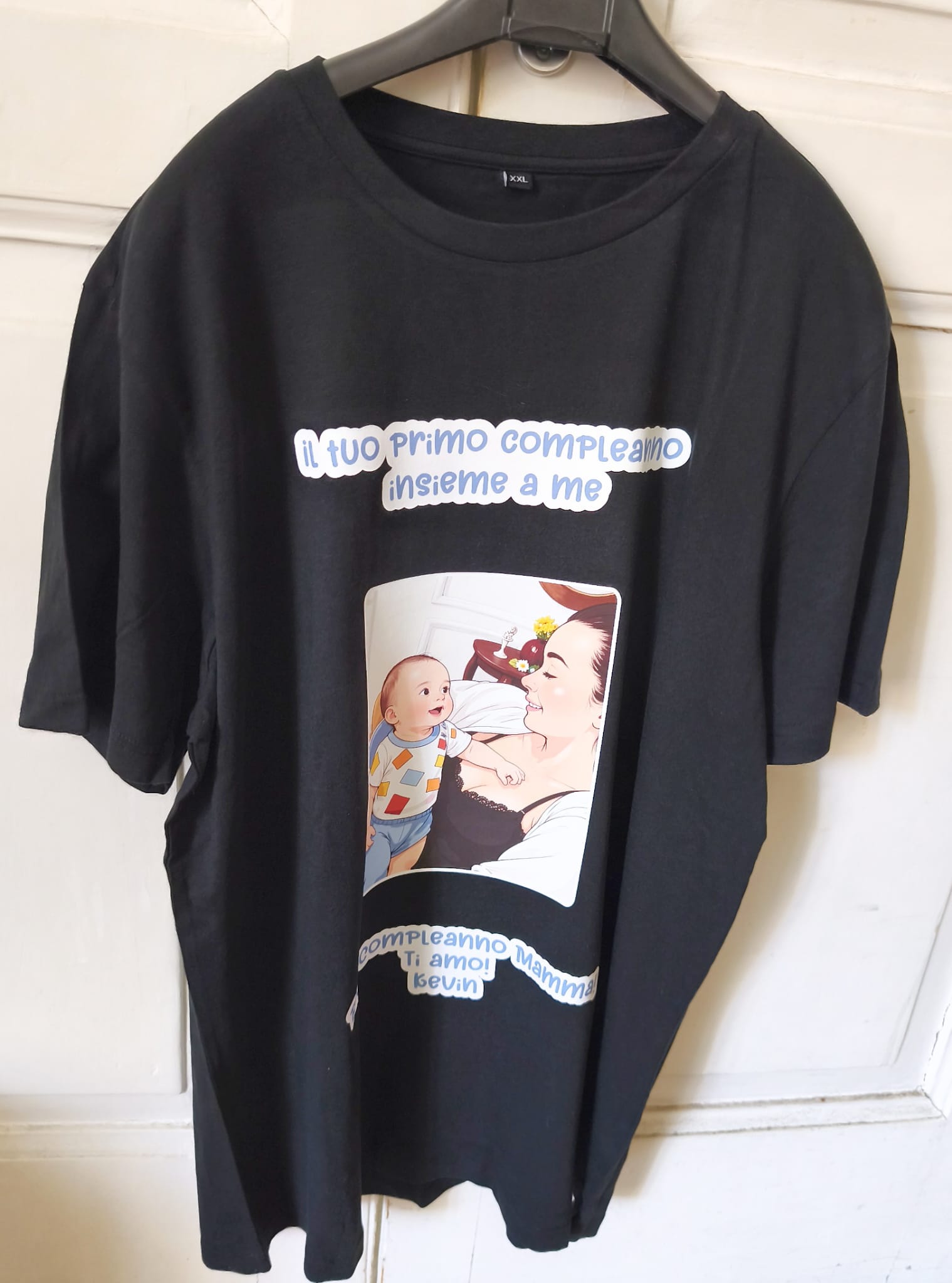 Black t-shirt with cartoon of mother and baby, Italian birthday message, on hanger indoors