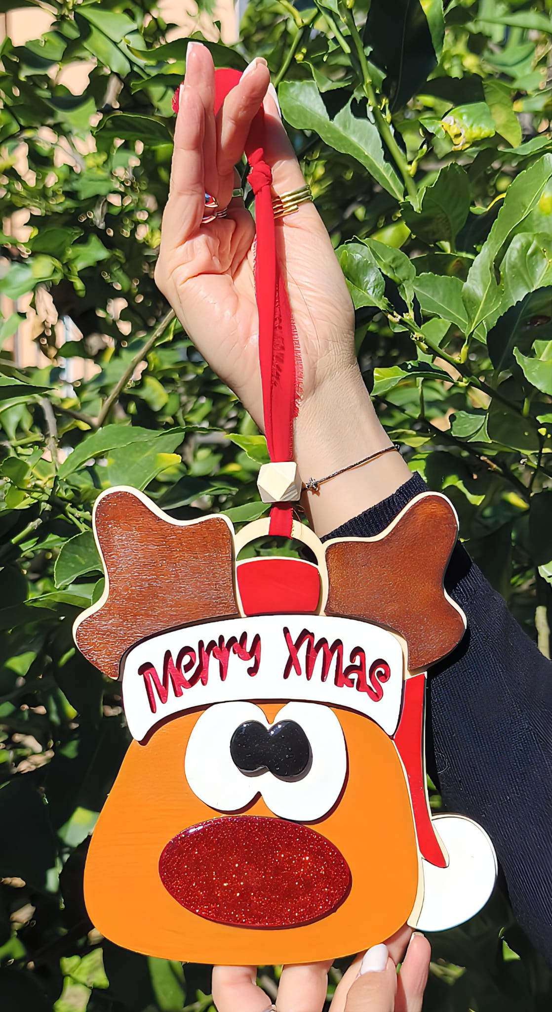 Hand holding Merry Xmas reindeer ornament with red glitter nose outdoors in greenery