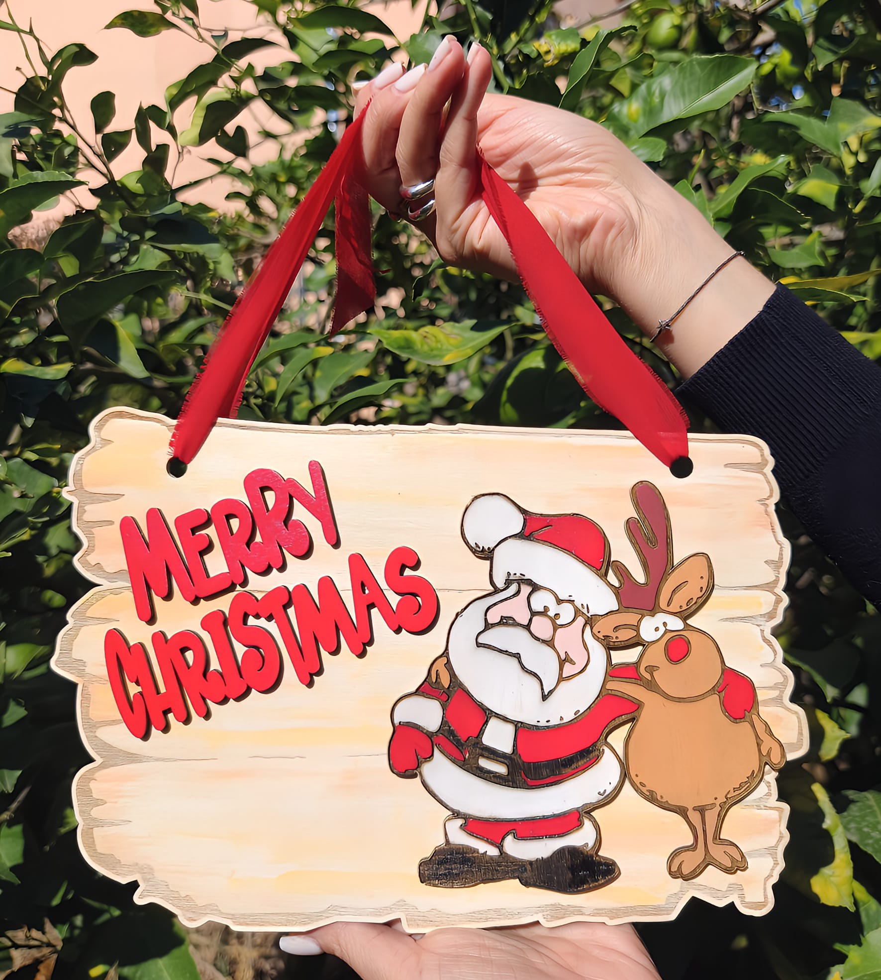 Hand holding Merry Christmas sign with cartoon Santa and reindeer, red ribbon, outdoor greenery