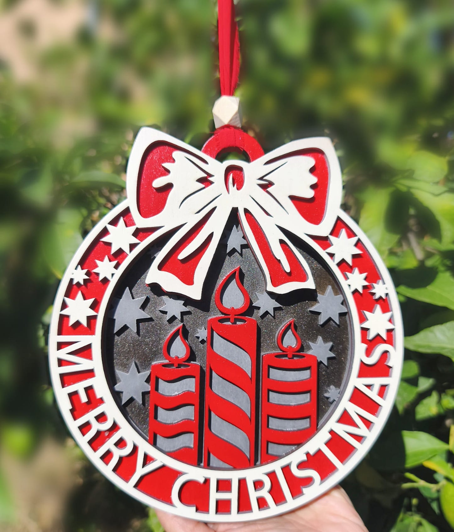 Red and white Merry Christmas ornament with candles, stars, and bow, hanging outdoors